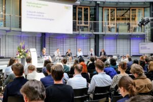 Blick ins Allianz Forum Berlin während der Podiumsdiskussion des UBi-Dialogforums 2026 mit Publikum und Bühne.