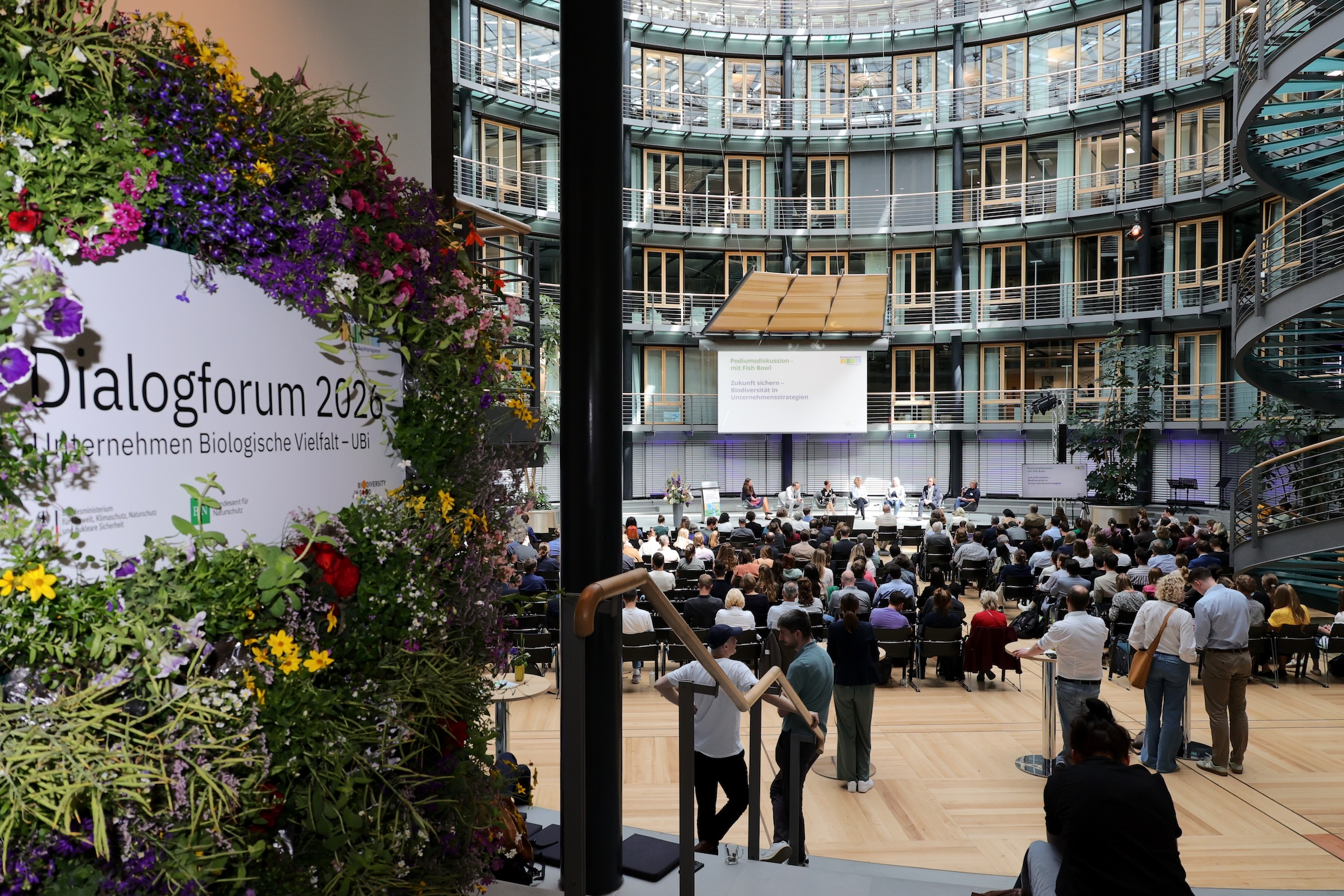 Blick in das Allianz Forum Berlin beim Dialogforum 2026 von „Unternehmen Biologische Vielfalt – UBi“ mit Bühne, Publikum und floralem Veranstaltungsaufsteller.