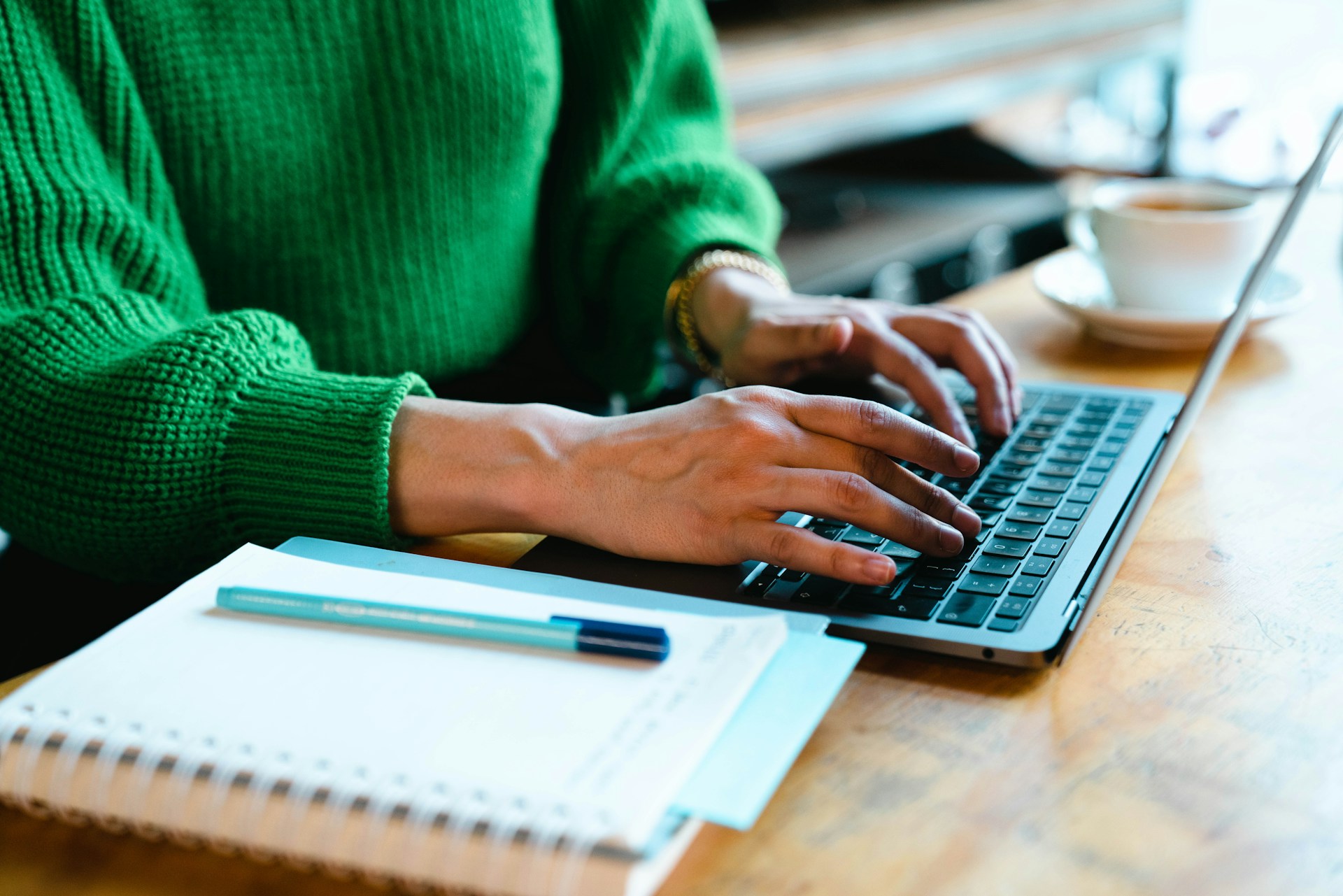 Person im grünen Pullover tippt an einem Laptop, daneben liegen Notizblock und Stift.